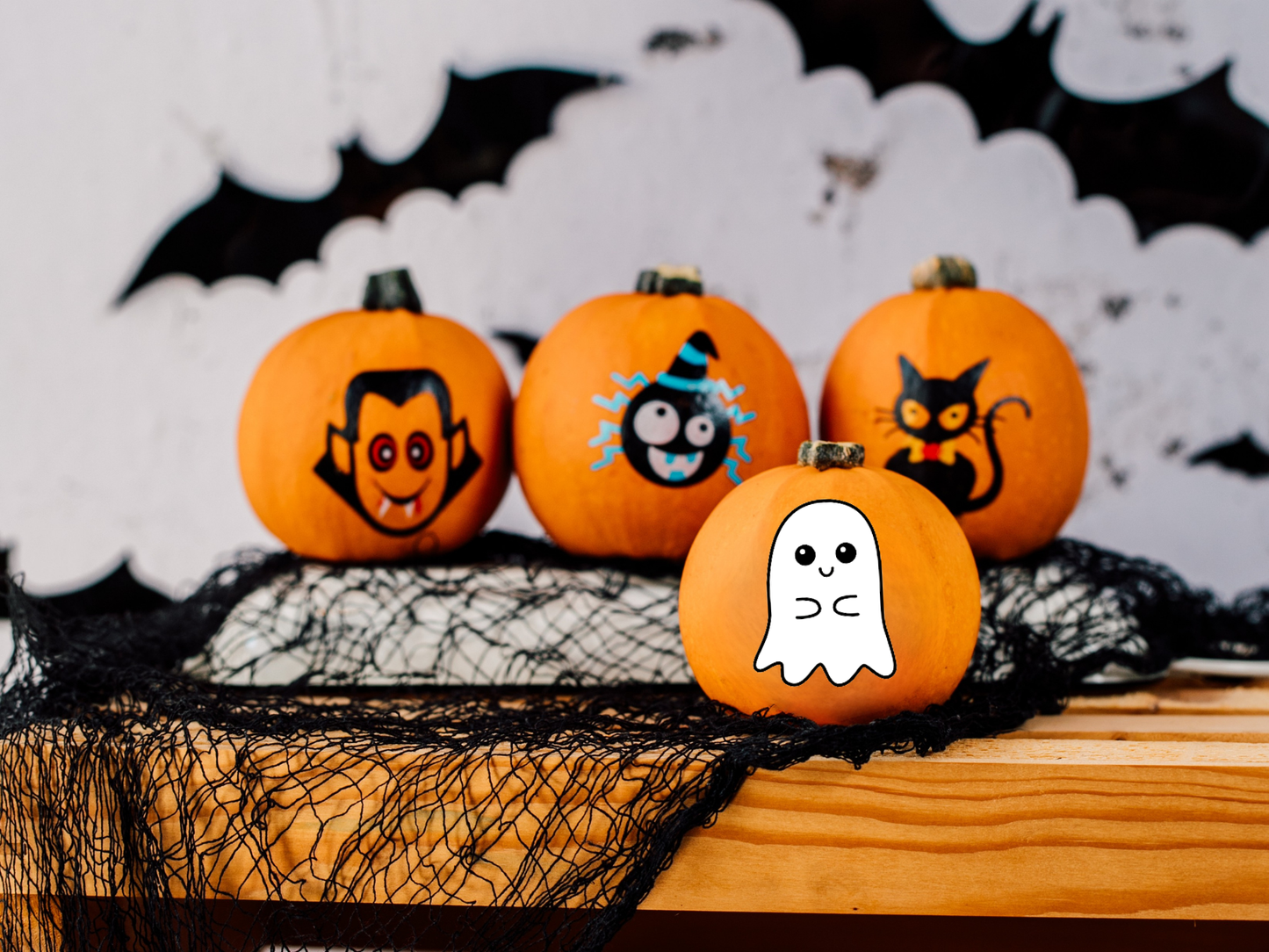 Decorative pumpkins with Halloween-themed faces on a wooden surface with black netting and bats in the background.