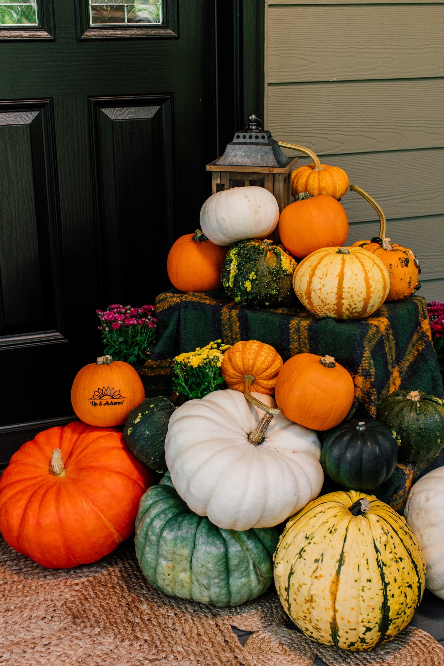 Stack of pumpkins and gourds on a porch with a lantern and flowers.