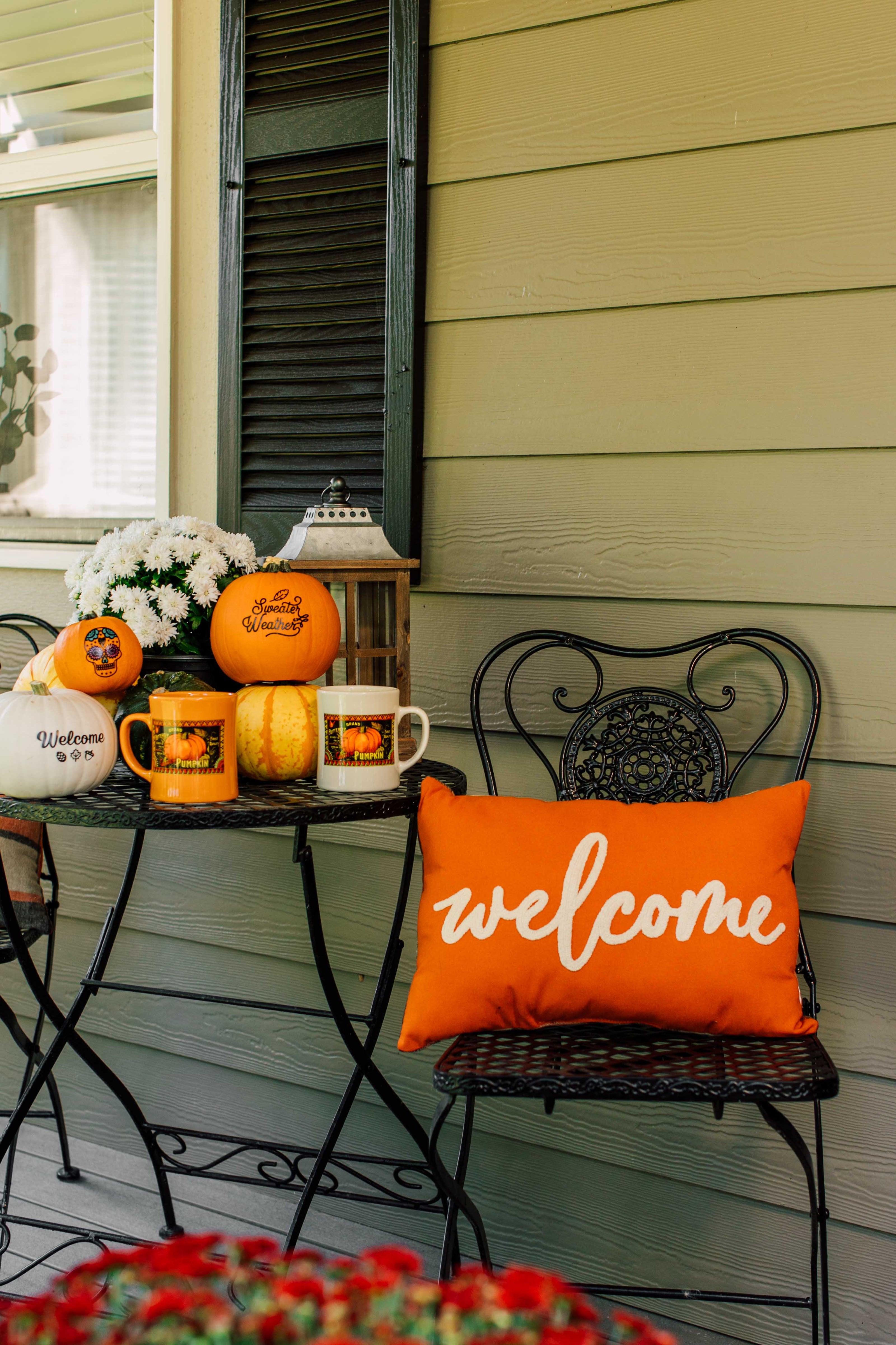 Outdoor setting with pumpkins, a 'welcome' pillow, and a small table on a porch.