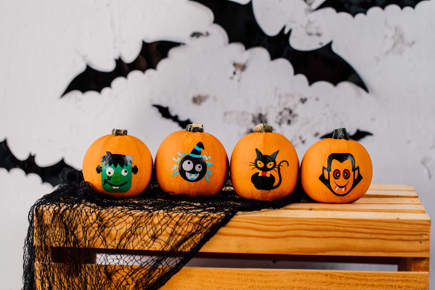 Four Halloween-themed pumpkins with face paint on a wooden surface with a bat and spider web background.