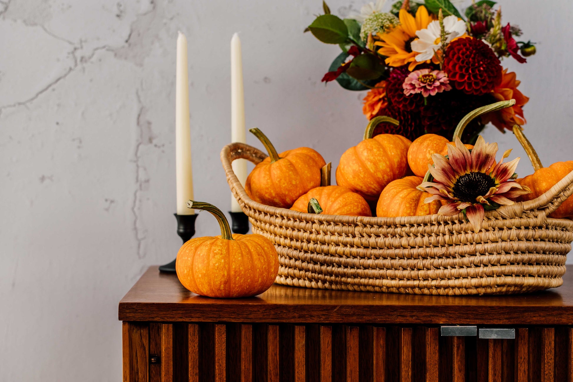 Basket of pumpkins with a candle and flowers on a wooden surface against a light gray wall.