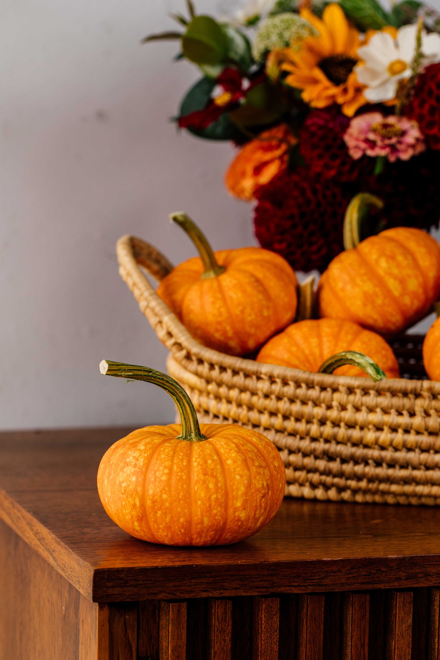 Small pumpkins on a wooden surface with a woven basket of flowers in the background.