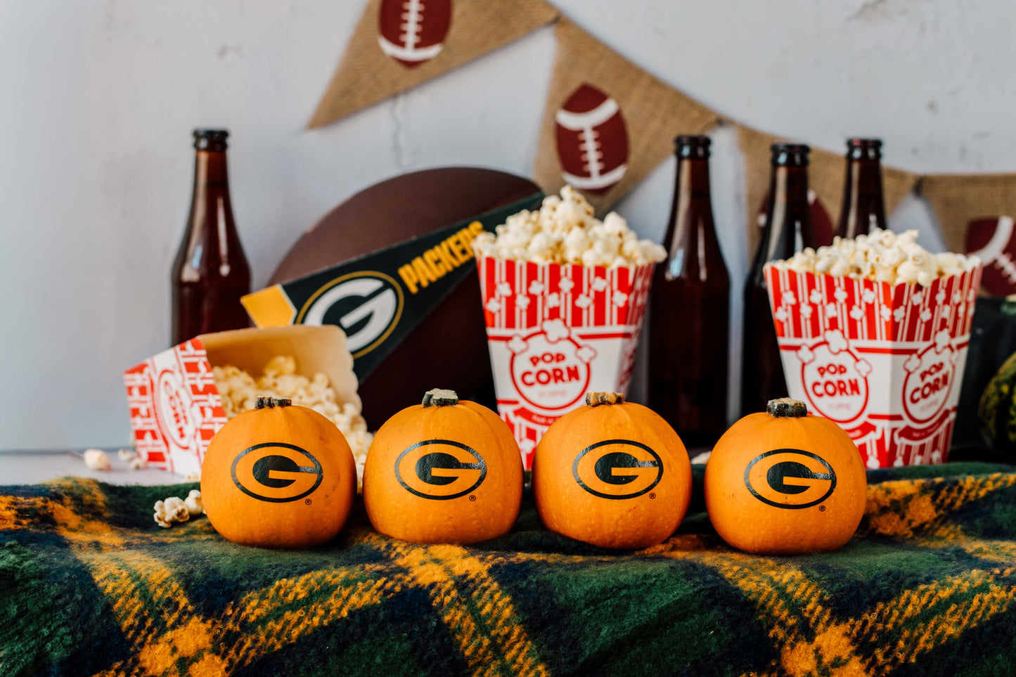 Pumpkins with Green Bay Packers logo, popcorn, and beer bottles on a plaid blanket.