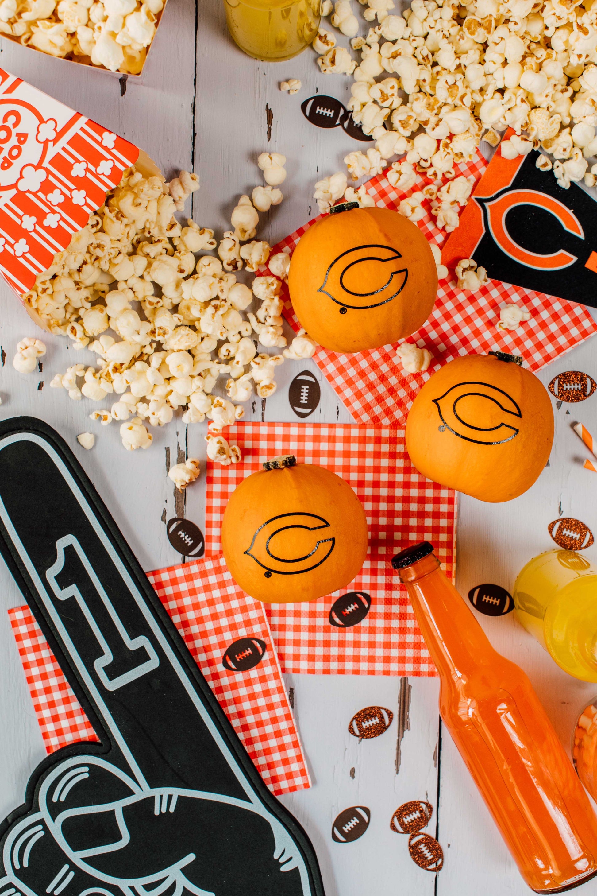 Orange pumpkins with black logos on a checkered cloth, surrounded by popcorn and a foam finger, on a light wooden surface.