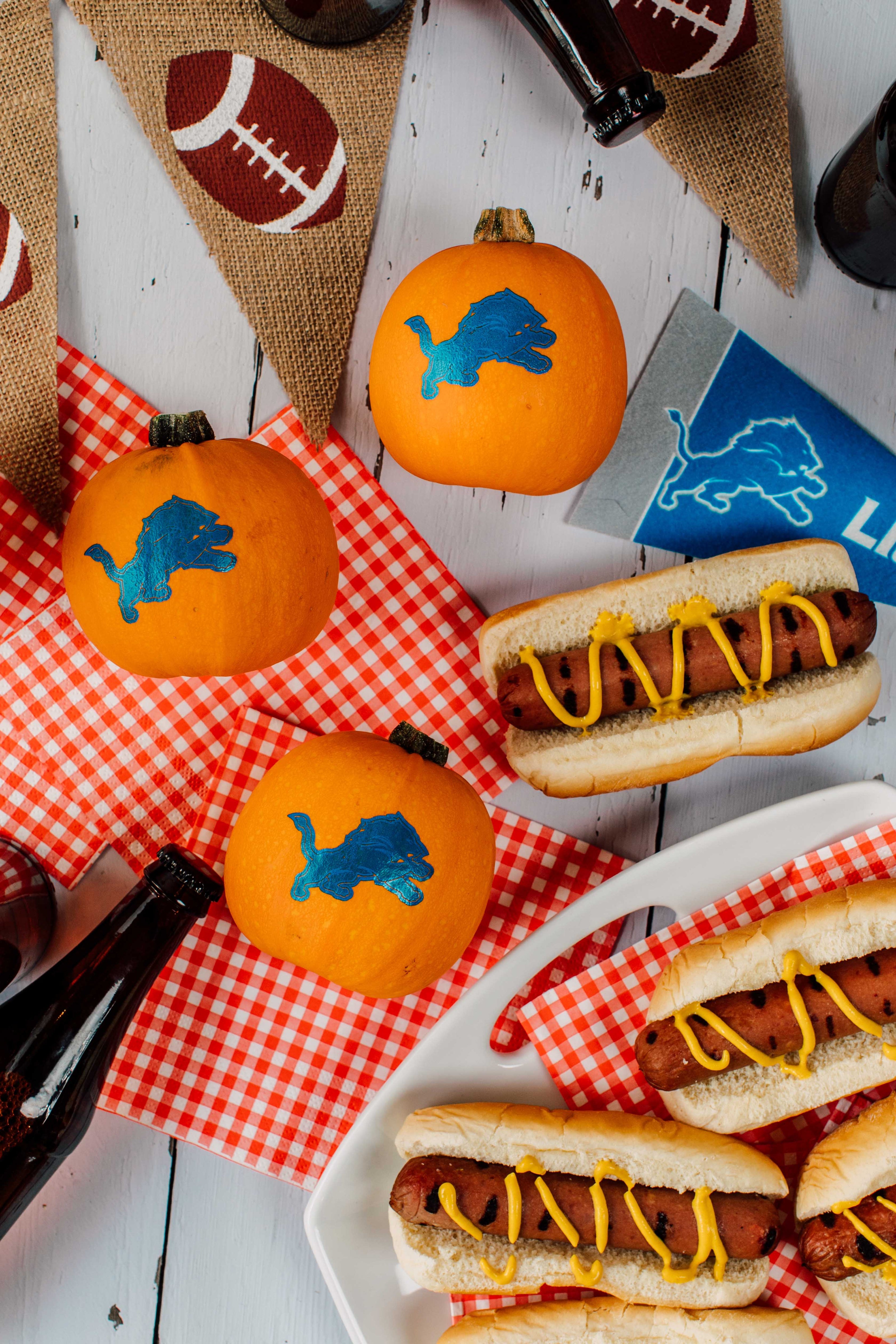 Hot dogs with mustard on a plate, surrounded by small pumpkins with blue logos and football-themed decorations.
