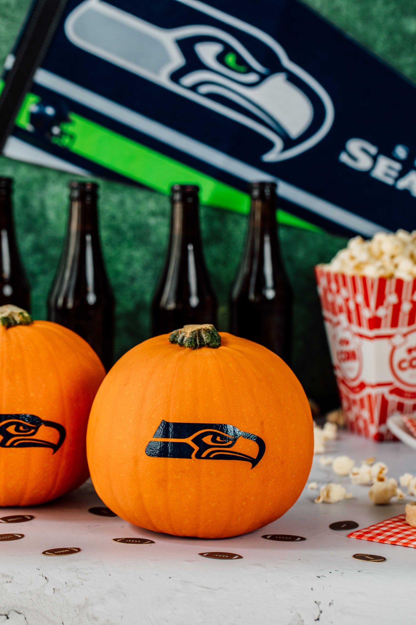 Two pumpkins with Seahawks logo, bottles, and popcorn on a table with a Seahawks banner in the background.