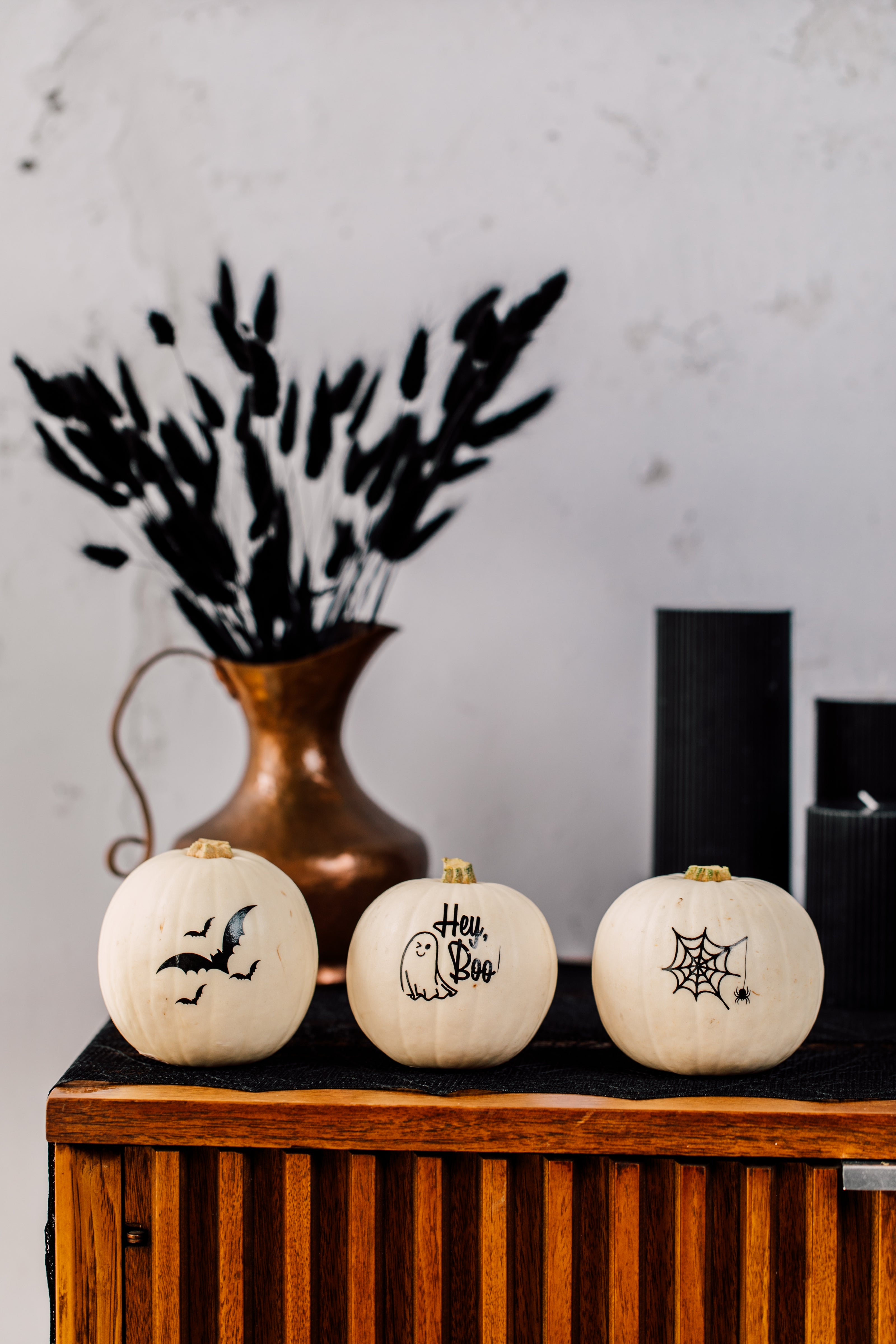 Three white pumpkins with Halloween designs on a wooden surface with a vase and candles in the background.
